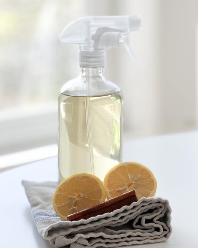 clear glass spray bottle with homemade lemon and cinnamon cleaning spray inside. Sitting on top of oranges, cinnamon, and towel