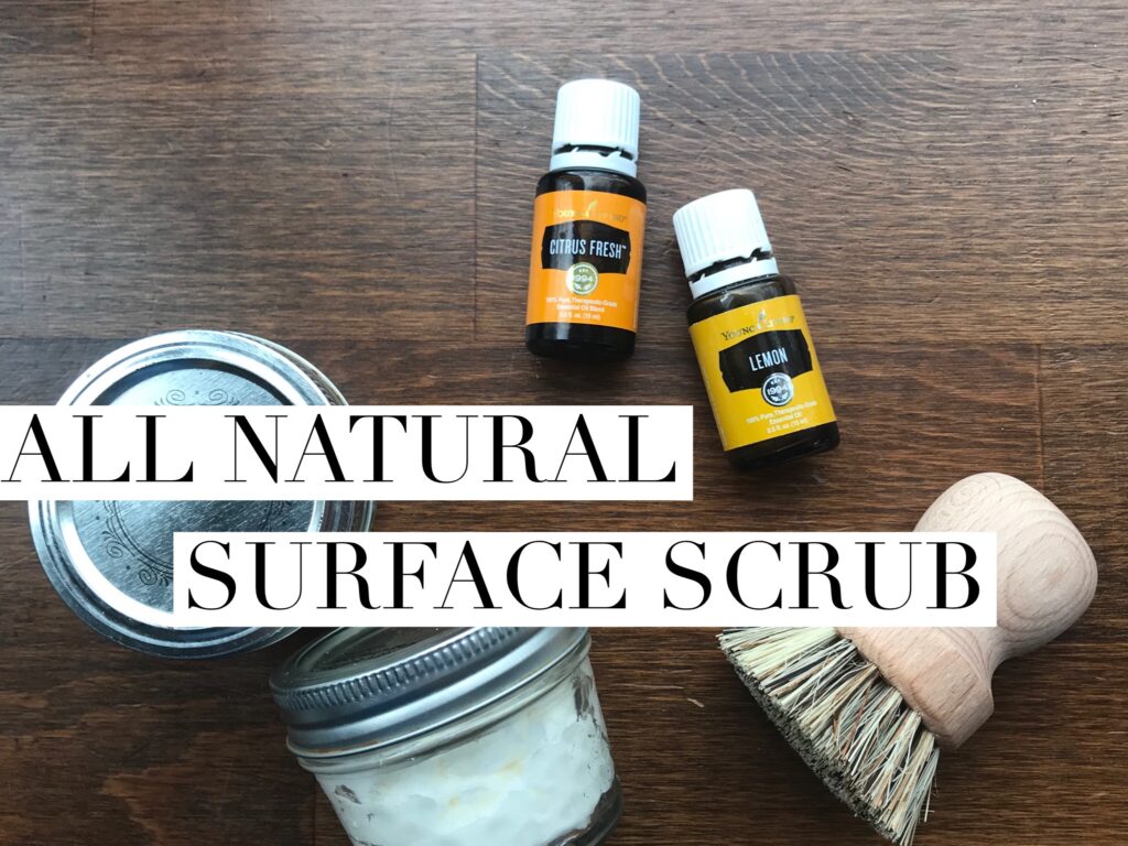 Essential oils, wood scrub brush, and glass jars filled with homemade cleaning scrub - text reads: All natural surface scrub
