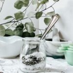 Thyme and Lavender fizzing bath salts in a glass jar with a metal spoon placed in the jar. On a white table with a white background and eucalyptus in the background.