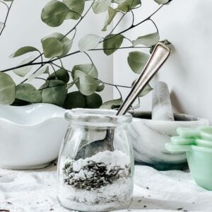 Thyme and Lavender fizzing bath salts in a glass jar with a metal spoon placed in the jar. On a white table with a white background and eucalyptus in the background.