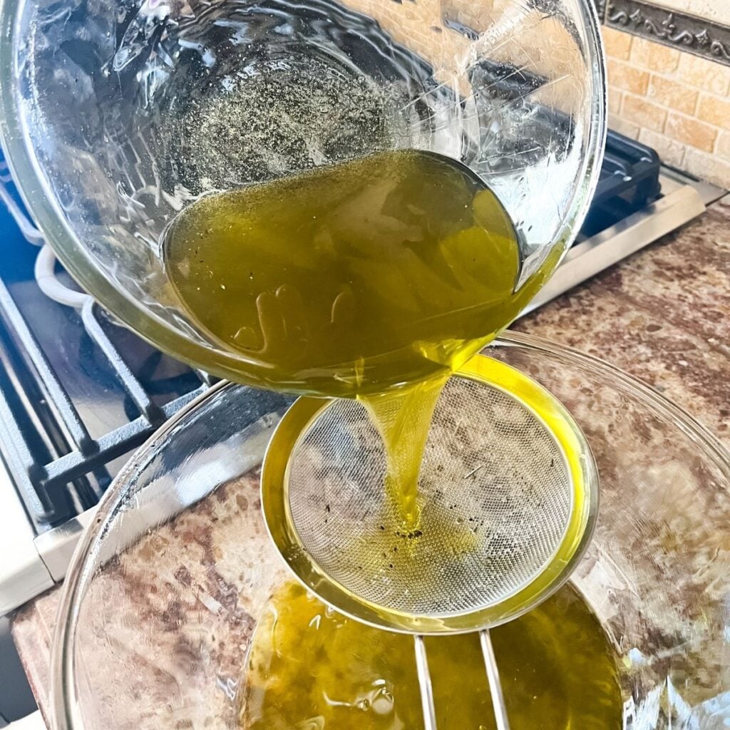 A bowl pouring the lavender-infused olive oil through a fine mesh sieve into a large glass bowl.