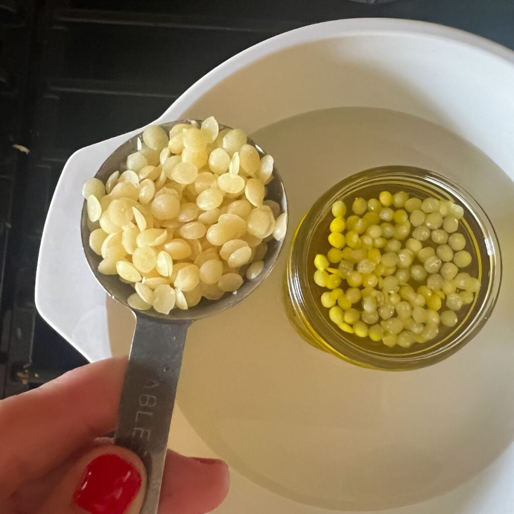 a hand holding a measuring spoon filled with beeswax pellets.
