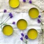 5 metal pots of lavender salve on a table with a white tablecloth surrounded by fresh lavender.